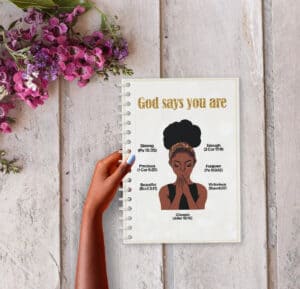 “An A5 wirobound notebook with a cover featuring the title ‘God says you are’ in gold lettering, accompanied by a praying woman illustration wearing a patterned headwrap. Surrounding the illustration are affirmations with corresponding Bible references: ‘Strong (Ps 18:35)’, ‘Enough (2 Cor 12:9)’, ‘Precious (1 Cor 6:20)’, ‘Forgiven (Ps 103:12)’, ‘Beautiful (Eccl 3:11)’, and ‘Victorious (Rom 8:37)’. The notebook is held against a rustic wooden background, adorned with vibrant purple flowers on the side.”