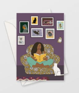 greeting card is shown with a white envelope. the card shows illustration of brown skinned woman reading a book called cat lady. She is sat cross legged in a comfy armchair with a white cat looking up at her and pictures of cats on the wall behind her. the wall is purple and the floor is dark brown.