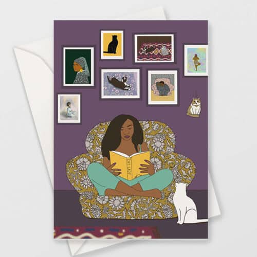 greeting card is shown with a white envelope. the card shows illustration of brown skinned woman reading a book called cat lady. She is sat cross legged in a comfy armchair with a white cat looking up at her and pictures of cats on the wall behind her. the wall is purple and the floor is dark brown.